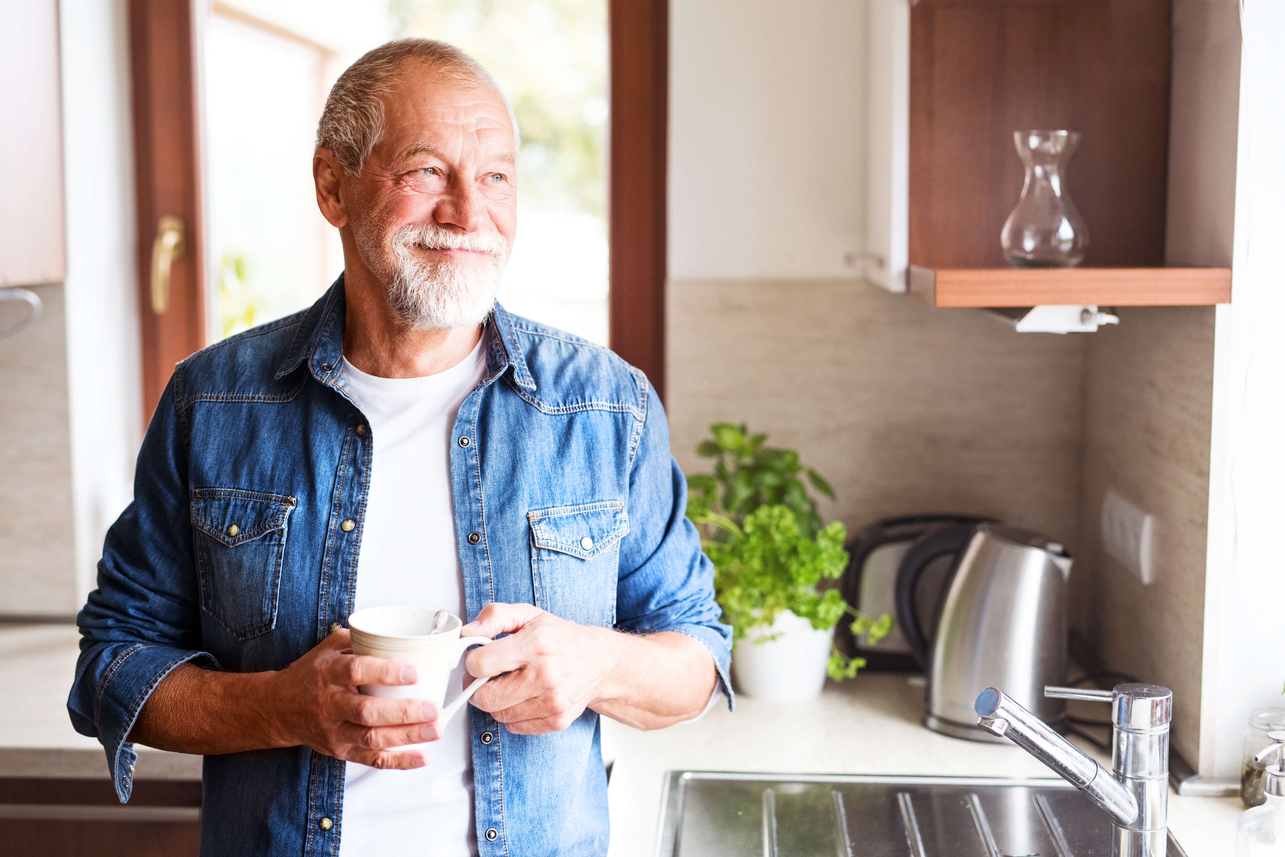 elder white male wearing a denim shirt holding coffee mug looking content out the window